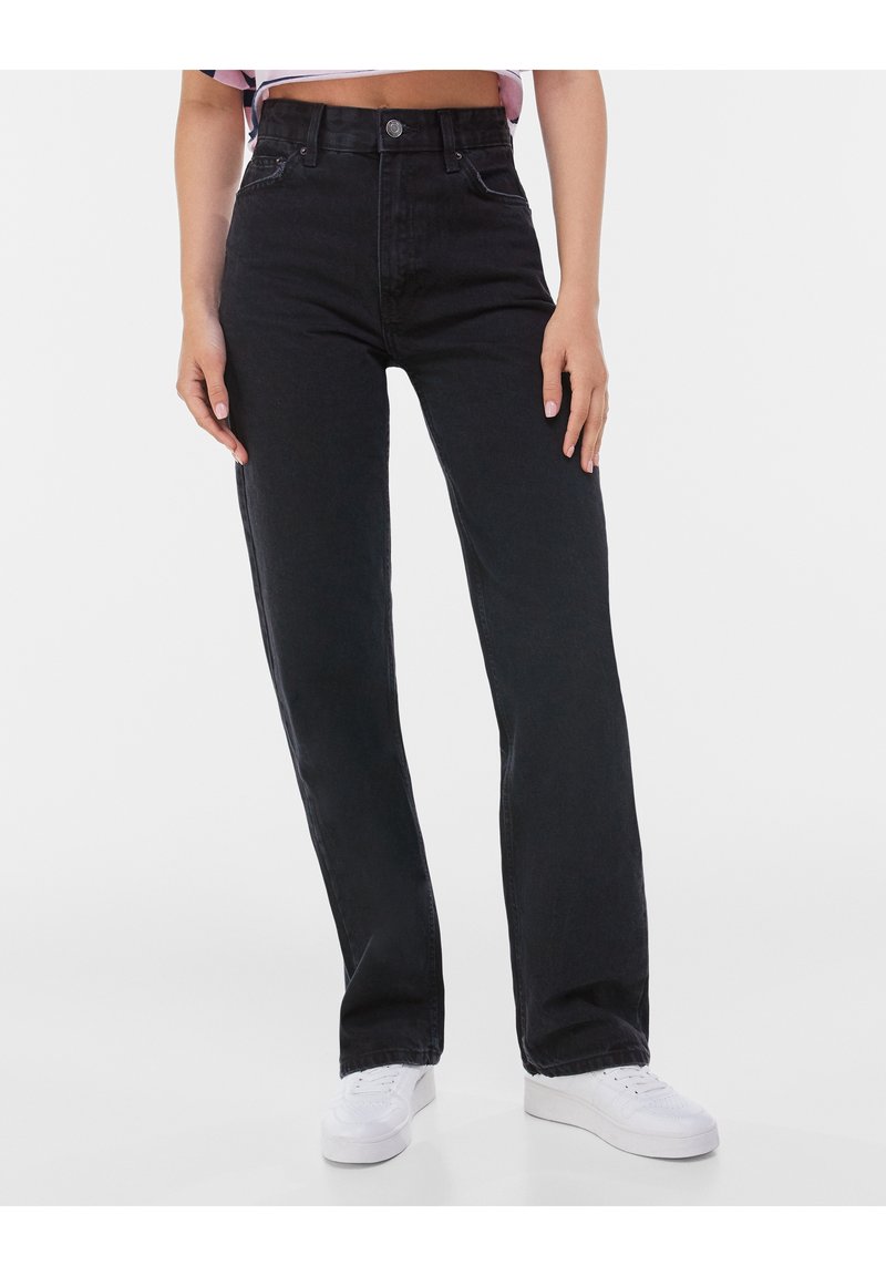 Personne portant un jean noir taille haute à jambe droite et des baskets blanches, crop top visible, debout devant un fond uni et clair.