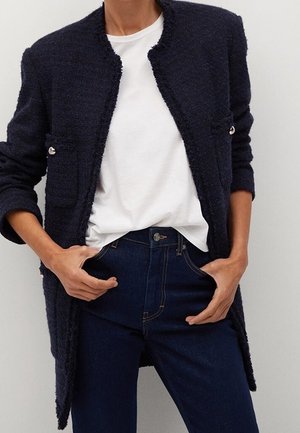 Femme portant un manteau texturé bleu marine foncé par-dessus un t-shirt blanc rentré dans un jean bleu foncé, mains reposant près des poches.
