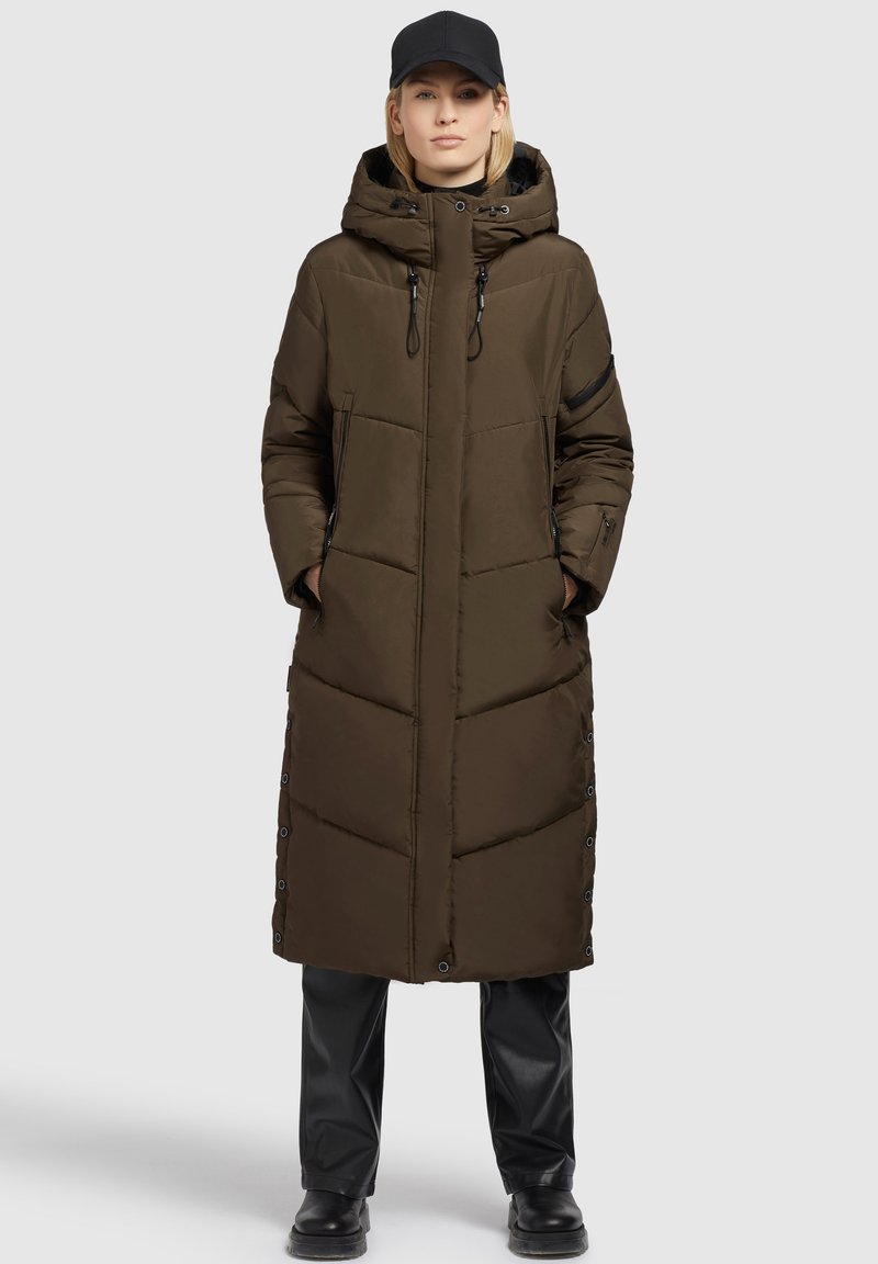 Frau trägt einen langen braunen gefütterten Mantel mit Kapuze, Frontreißverschluss, seitlichen Druckknöpfen, schwarze Hose, schwarze Stiefel und eine schwarze Baseballkappe.