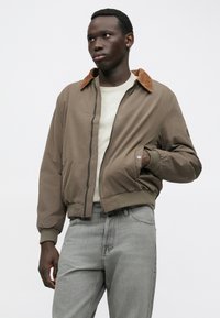 Νεαρός άνδρας φορώντας καφέ τζάκετ bomber με ταμπά κολάρο από καπιτονέ, λευκό πουκάμισο, ανοιχτό γκρί τζιν, που στέκεται με το ένα χέρι στην τσέπη.