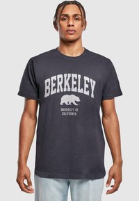 Tmavě šedé tričko vyrobené z bavlny má na sobě potisk "BERKELEY" a grafiku medvěda v světle šedé barvě, s nápisem "UNIVERSITY OF CALIFORNIA" pod tím.