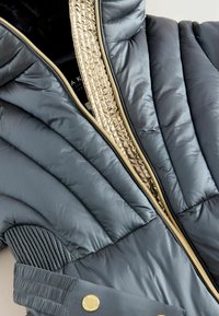 Chaqueta acolchada en gris oscuro con un cierre dorado, que presenta detalles texturizados a lo largo del cierre y un material acanalado en los paneles laterales.