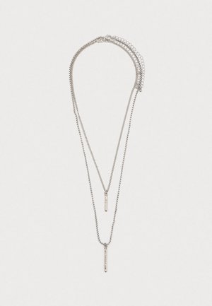 Collier en argent superposé avec deux pendentifs en barres verticales minces sur des chaînes délicates sur fond blanc.