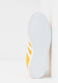 Gul sneaker med vita ränder, texturerad gummisula och mjukt tygöverkänsla. Designen har en låg siluett och noggrant sydda detaljer.