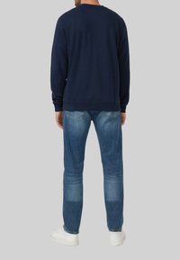 Uomo in piedi di spalle, indossando una felpa blu navy, jeans blu e sneaker bianche su uno sfondo grigio.