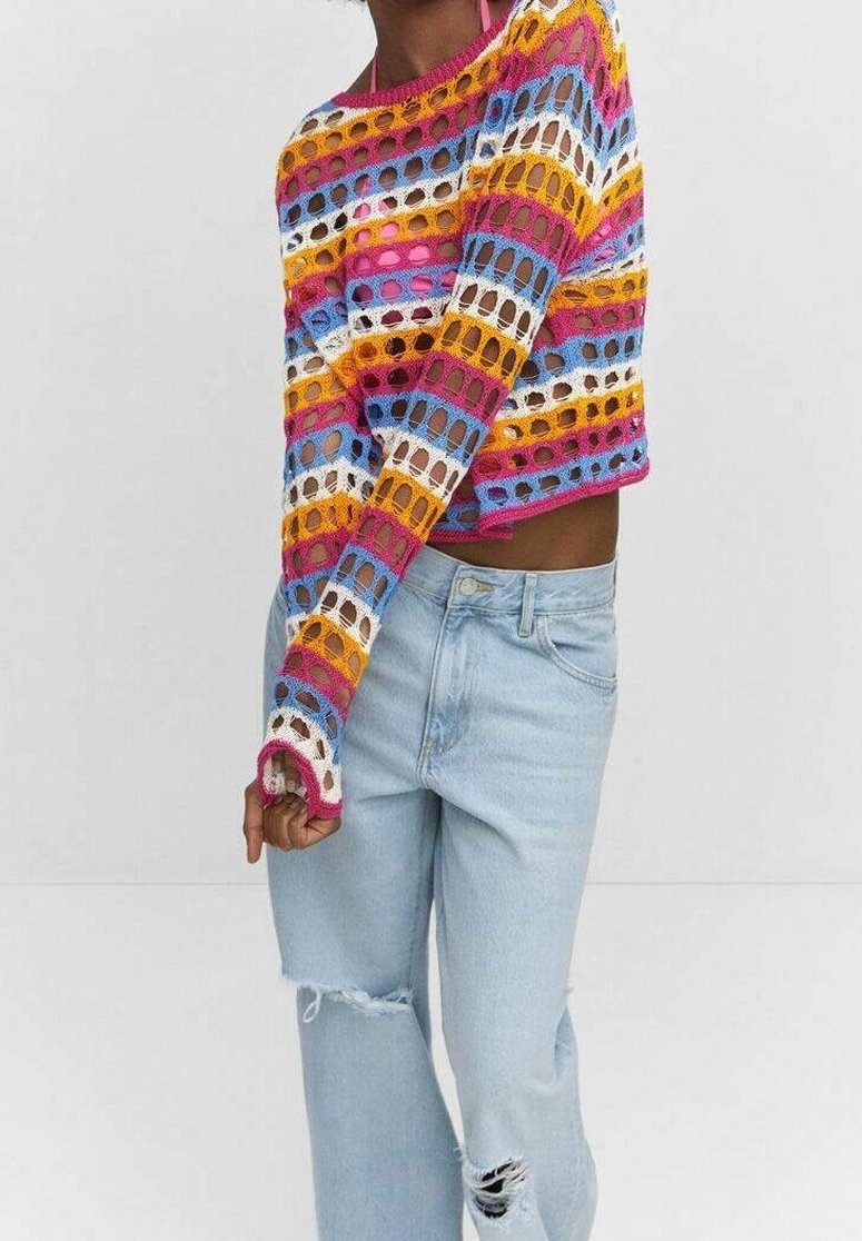 Personne portant un pull coloré à rayures en maille ouverte et un jean bleu clair déchiré, debout devant un fond blanc uni.