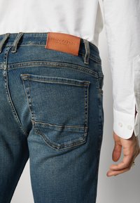 Dark blue denim jeans with a leather label on the waistband. Features traditional five-pocket styling and visible stitching details.