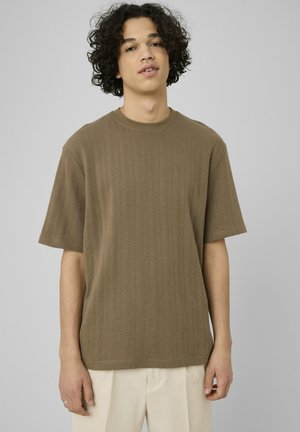 Jeune homme aux cheveux bouclés portant une chemise texturée à manches courtes vert olive et un pantalon beige clair sur un fond gris uni.