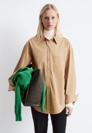 Beige Hemd mit Knöpfen und einer Brusttasche, darüber getragen ein weißer Rollkragenpullover. Hält einen grünen Strickschal und eine schwarze Ledertasche.