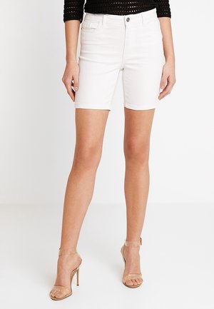 Kvinde iført hvide shorts ned til knæene, sort cropped top og klare højhælede sandaler, stående med benene let spredt.