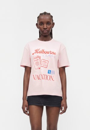 Kobieta z zaplecionymi włosami, ubrana w różowy t-shirt z czerwonymi stemplami podróżniczymi i napisem "Melbourne Australia VACATION" oraz czarną mini spódniczkę.