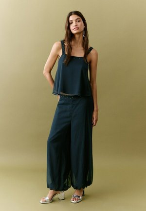 Kobieta ubrana w ciemnoturkusowy, bezrękawnik z luźnym krojem oraz pasujące szerokie spodnie i srebrne sandały na obcasie blokowym na tle jednolitego beżowego tła.