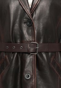 Tmavohnedý kožený kabát s veľkými čiernymi gombíkmi, zodpovedajúcim koženým opaskom s kovovou prackou a šikmými prednými vreckami.