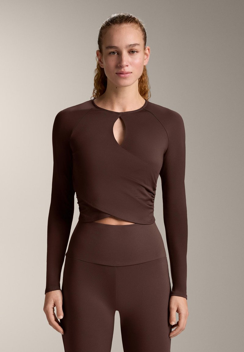 Jonge vrouw staat met het gezicht naar voren en draagt een nauwsluitend, donkerbruin, lang mouw top met keyhole uitsparing en bijpassende hooggetailleerde leggings.