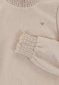 Lichtbeige lang mouwtop met geribbelde textuur, elastische manchetten en een kleine roze hartborduring dicht bij de halslijn.