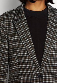 Blazer a quadri in marrone, nero e bianco con rever a punta, chiusura con un bottone e una texture morbida. Indossato sopra un maglione scuro.