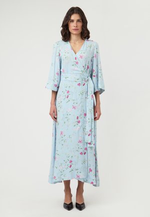 Kobieta w jasnoniebieskiej, kwiecistej sukience kopertowej z rękawami do połowy przedramienia i czarnych szpilkach z zaostrzonymi noskami, stojąca na jednolitym tle.