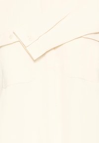 Close-up of an off-white long-sleeve shirt with visible white buttons on cuff and two chest pockets with stitching details.