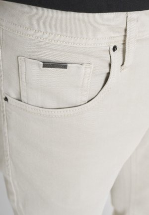 Nahaufnahme einer beigen Hosentasche mit einem kleinen rechteckigen Metallschild, das an der Innenkante der Tasche befestigt ist.