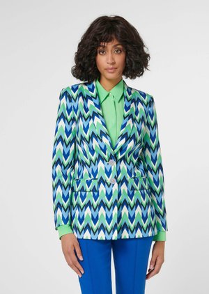 Frau mit lockigem dunklem Haar, die einen Blazer mit blau-grün-weißem Zickzackmuster über einem grünen Hemd und blauen Hosen trägt, stehend vor weißem Hintergrund.