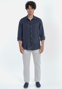 Donkerblauwe linnen overhemd met een knoopsluiting, opgerolde mouwen, gedragen met lichtbeige linnen broek en marineblauwe schoenen. Effen achtergrond.