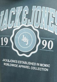 Tume roheline puuvillane T-särk, millel on helesinised trükitud tähed, ringikujuline logo, millel on "J&J," ja "JACK&JONES" asutamise üksikasjad.