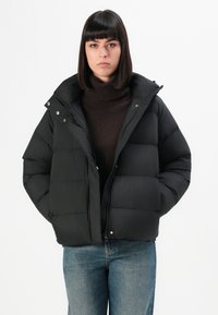 Black puffer jacket with a zip front, snap buttons, and a wide collar. Features quilted sections and a relaxed fit. Worn over a brown turtleneck.