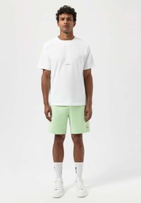 Bílá bavlněná tričko s logem na přední straně, spárované se světle zelenými šortkami a bílými teniskami. Model nosí bílé ponožky s černým brandingem.