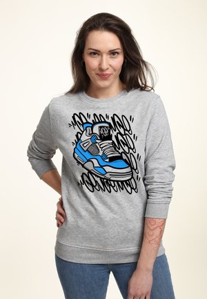 Kobieta z długimi ciemnymi włosami, ubrana w szarą bluzę z grafiką niebieskiego i czarnego sneakersa oraz niebieskie dżinsy, stojąca z ręką na biodrze.