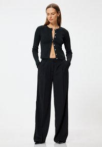 Zwarte geribbelde cardigan met knoopsluiting en lange mouwen, gecombineerd met hoge zwarte broeken. Glad materiaal, casual pasvorm, minimale accenten.