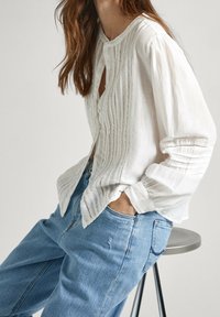 Blusa di lino bianco con delicati ricami verticali e bottoni, abbinata a jeans di denim azzurro. Vestibilità comoda e maniche arrotolate.