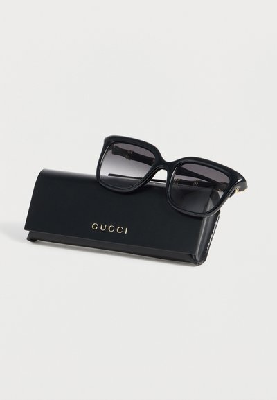 Μαύρα γυαλιά ηλίου Gucci με σκούρους φακούς που ακουμπούν σε μαύρη θήκη Gucci πάνω σε λευκή επιφάνεια.
