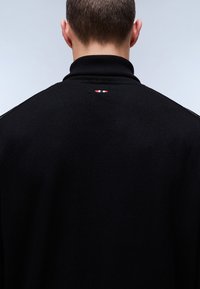 Zwarte gebreide kleding met een hoge kraag. Heeft een klein rood, wit en blauw logo op de achterkant. Soepele textuur en aansluitend ontwerp.