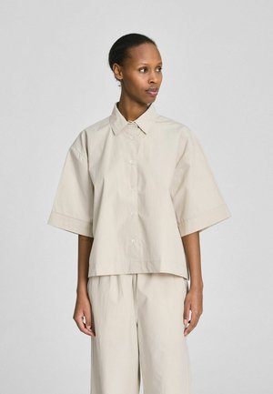 Vrouw draagt een losse beige blouse met korte mouwen en knopen, en bijpassende wijde broekspijpen, kijkt lichtjes opzij tegen een effen achtergrond.