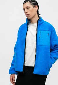Niebieska polarowa kurtka z czarnym zamkiem błyskawicznym, z zamykaną na zamek przednią kieszenią i czarnym logo. Gładki, teksturowany materiał i luźny krój.