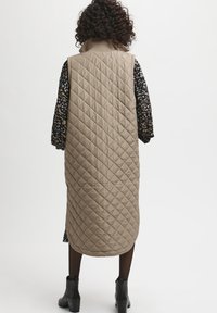 Frau mit lockigem Haar trägt eine lange, beigefarbene, gesteppte Weste über einem gemusterten Kleid und schwarzen Stiefeletten und steht vor einem weißen Hintergrund.