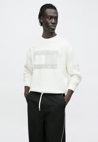 Κρεμ φούτερ με γκρι υφασμένη λογότυπο που αναγράφει "TOMMY" και "JEANS" στο μπροστινό μέρος, συνδυασμένο με μαύρο παντελόνι με κορδόνι.