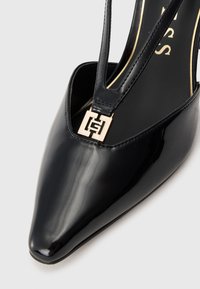Pantofii cu toc înalt din piele lacuită neagră, cu vârf ascuțit, prezentând un design cu decupaje și un accent cu logo pătrat auriu pe curea.