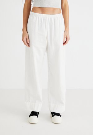 Persoon die losse witte broek draagt met elastische tailleband en zwart-witte sneakers, staand tegen een witte achtergrond.
