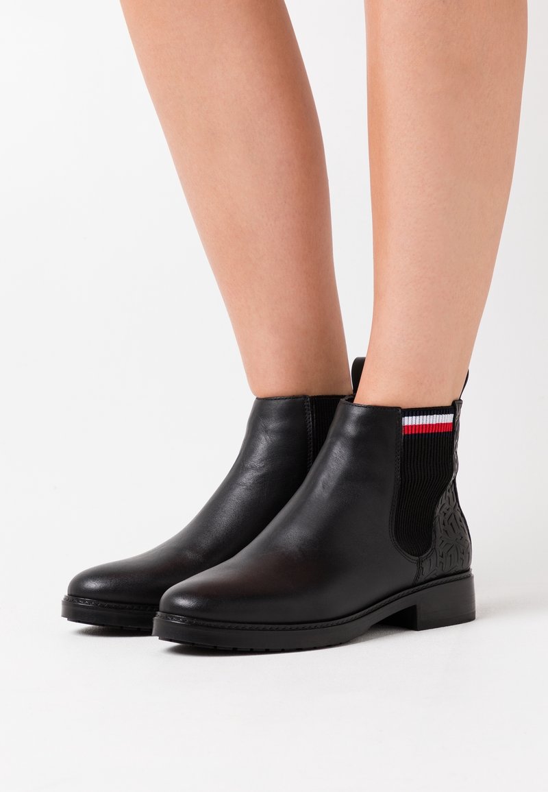 Bottines en cuir noir avec côtés élastiques côtelés ornés de rayures rouges, blanches et bleues, portées sur des jambes nues sur fond blanc.