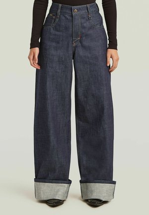 Person trägt weit geschnittene dunkelblaue Jeans mit großen Umschlägen, kombiniert mit schwarzen spitzen Schuhen und einem schwarzen Langarm-Top.