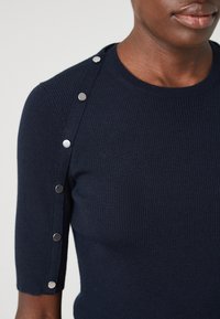 Tmavě modrý top z žebrovaného úpletu s kulatým výstřihem, polovičními rukávy a stříbrnými druky podél švu na rameni. Hladká textura.