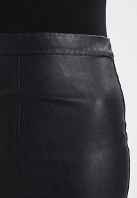 Jupe en cuir noir avec une texture lisse, un design ajusté et une large taille. Présente des coutures sur les côtés pour plus de structure.