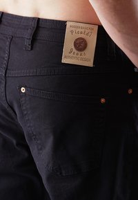Schwarze Jeans mit einem Lederetikett, das "Picaldi Jeans Authentic Design" liest. Merkmale sind Gesäßtaschen mit Kupferbeschlägen.