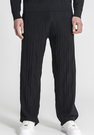 Man draagt zwarte textuurggebreide broek en witte sneakers, staand voor een effen lichte achtergrond.