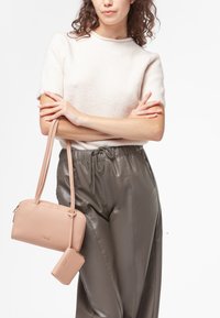 Borsa a tracolla in pelle rosa chiaro con superficie testurizzata, chiusura con zip e pochette piccola staccabile, abbinata a pantaloni grigi a pieghe e a un maglione beige.