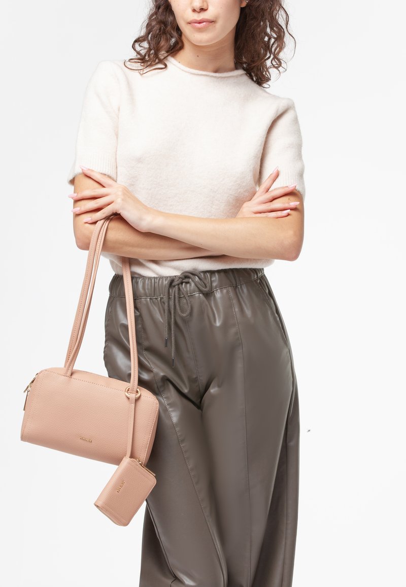Borsa a tracolla in pelle rosa chiaro con superficie testurizzata, chiusura con zip e pochette piccola staccabile, abbinata a pantaloni grigi a pieghe e a un maglione beige.