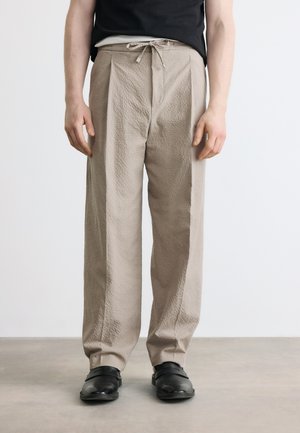 Mężczyzna ubrany w beżowe, teksturowane spodnie ze ściągaczem w pasie i czarne buty wsuwane, stojący na tle jednolitego jasnego tła.