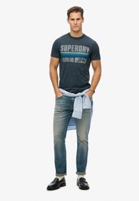 Camiseta gráfica azul marino con el texto "SUPERDRY", jeans de mezclilla azul claro, una camisa azul claro atada a la cintura y mocasines negros.