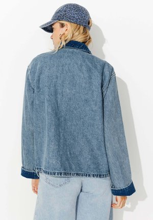 Femme portant une veste en denim bleu et un jean clair en denim, avec une casquette de baseball à imprimé léopard, vue de dos sur fond blanc.